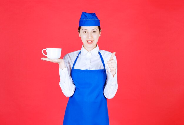 Cameriera della donna in uniforme che tiene una tazza e che mostra pollice su sulla parete rossa.