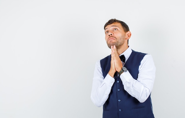 Cameriere appoggiando il mento sulle mani nel gesto di preghiera in camicia, gilet e guardando speranzoso, vista frontale.