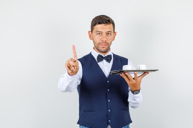 Cameriere che tiene tazze nel vassoio con gesto di attesa in camicia, gilet, farfallino, vista frontale.