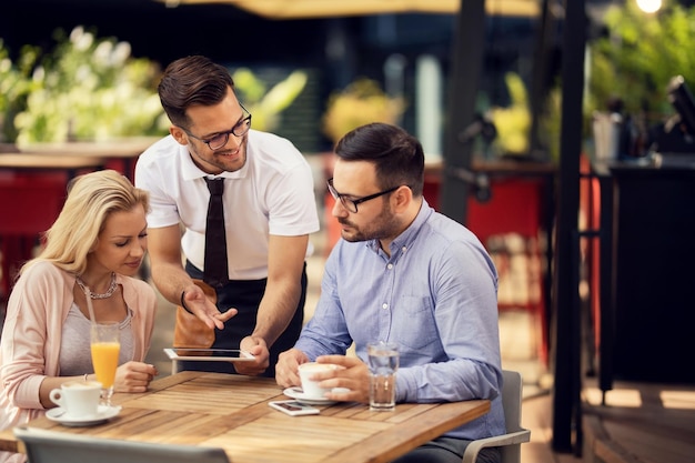 Cameriere felice che parla con gli ospiti mentre usa la tavoletta digitale e mostra loro il menu in un ristorante