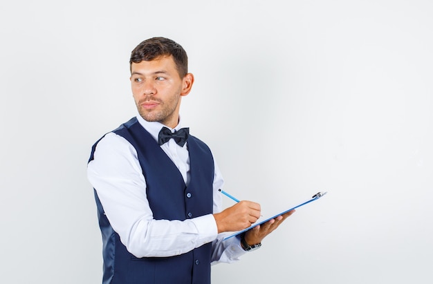 Cameriere in camicia, giubbotto guardando indietro mentre prende appunti negli appunti e sembra pensieroso, vista frontale.