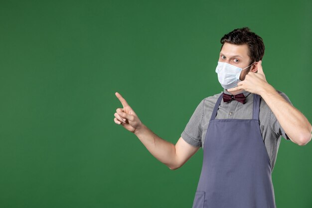 cameriere serio in uniforme con maschera medica e rivolto verso l'alto sul lato destro facendomi gesto di chiamata su sfondo verde