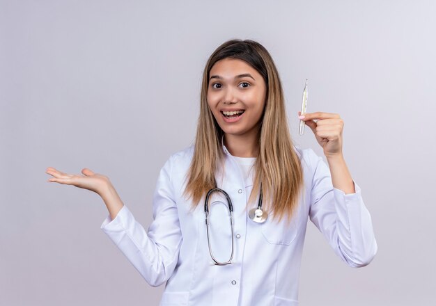 Camice bianco da portare del medico della giovane bella donna con il termometro della tenuta dello stetoscopio che sembra presentazione sicura con il braccio oh la sua mano