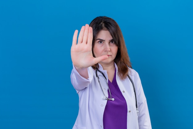 Camice d'uso di medico invecchiato mezzo e con lo stetoscopio con la mano aperta che fa il fanale di arresto con l'espressione seria e sicura