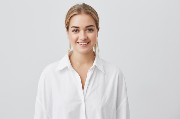 Camicia bianca d'uso della bella giovane donna allegra con capelli biondi che sorride piacevolmente mentre ricevendo alcune notizie positive. Ragazza graziosa che osserva con il sorriso allegro