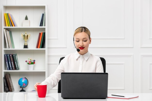 Camicia da ufficio ragazza bionda carina servizio clienti con auricolare e computer in possesso di una tazza rossa
