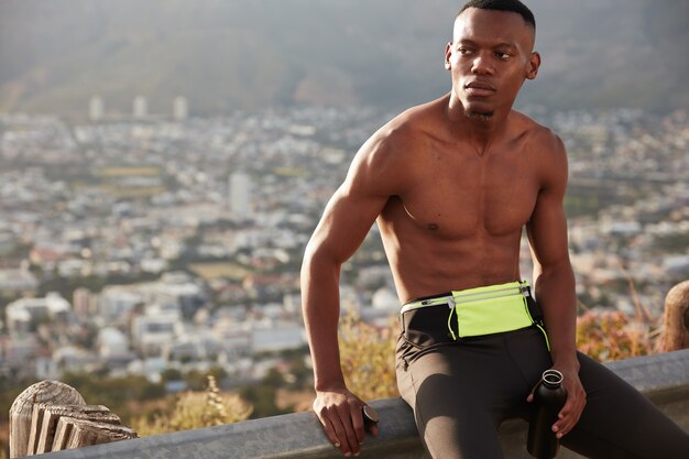 Campagna sano stile di vita attivo concetto. Uomo etnico concentrato con corpo muscoloso, tiene la bottiglia sportiva piena d'acqua, si prende una pausa dopo lo sprint, pronto per correre la maratona, fa jogging all'esterno