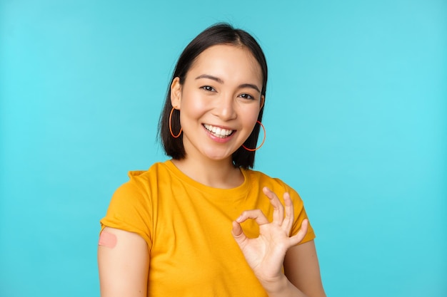 Campagna vaccinale da covid19 Ragazza asiatica in buona salute felice che mostra segno ok e cerotto sorridente sulla spalla da sfondo blu vaccinazione coronavirus