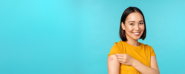 Campagna vaccinale di covid19 Giovane bella donna asiatica in buona salute che mostra la spalla con il concetto di vaccinazione in bandaid in piedi su sfondo blu