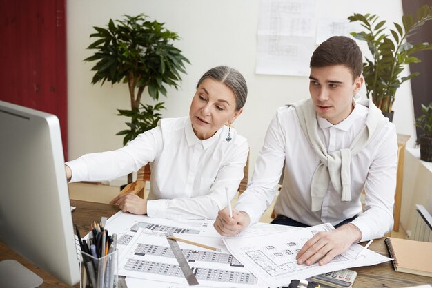 Candid colpo di attraente femmina matura in via di sviluppo progetto di costruzione in ufficio leggero, lavorando insieme al suo giovane collega maschio, utilizzando il sistema CAD su computer generico, indicando lo schermo