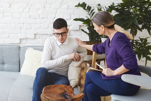 Candid colpo di psicoterapeuta professionista donna vestita casualmente sulla cinquantina che tocca il suo giovane paziente maschio a spalla mentre ha una sessione di consulenza, esprimendo simpatia e sostegno