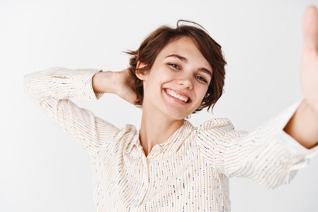 Candida donna felice che posa per selfie tenendo lo smartphone e scattare foto di se stessa con un sorriso allegro in piedi spensierato su sfondo bianco