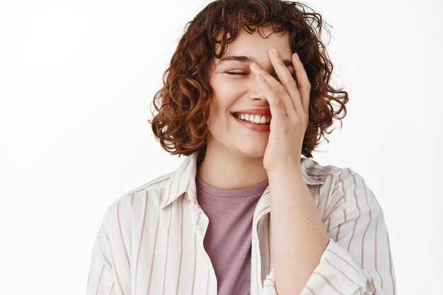 Candida donna felice con un bel sorriso, che copre il viso con la mano timida, arrossendo mentre ride e si diverte, in piedi rilassata su sfondo bianco