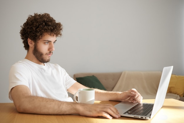 Candido colpo di bel giovane scapolo fiducioso con la barba folta che si rilassa a casa utilizzando la connessione Internet wireless ad alta velocità sul computer portatile, navigando su siti Web e bevendo caffè dopo il lavoro