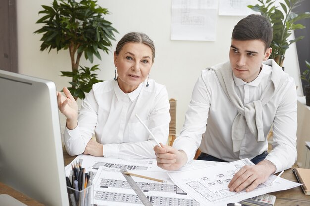 Candido colpo di elegante esperto architetto femmina senior dai capelli grigi che punta allo schermo del computer, spiegando il punto di vista al suo giovane collega maschio mentre lavorava insieme su un nuovo progetto abitativo,