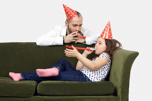 Candido ritratto di adorabile bambina graziosa che gode della festa di compleanno a casa, sdraiato sul divano e soffiando il corno della festa insieme al suo giovane padre con la barba lunga