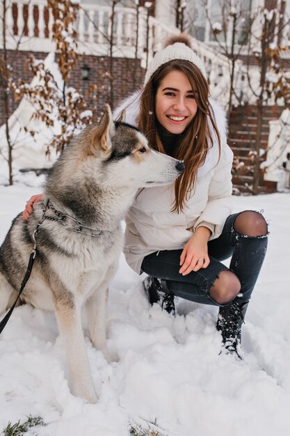 Cane fedele che guarda lontano nel giorno di inverno mentre ridendo donna in giacca bianca accarezzandolo. Spettacolare signora europea in jeans in posa con husky su terreno innevato.