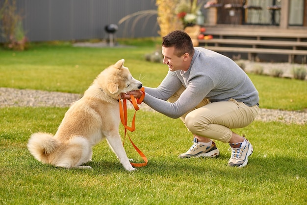 Cane intelligente. Lateralmente alla telecamera di un gioioso giovane adulto accovacciato che tocca un cane amichevole e intelligente con il guinzaglio vicino alla casa di campagna il giorno d'autunno