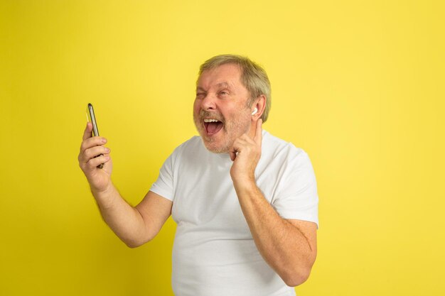 Cantando con auricolare e smartphone. Ritratto di uomo caucasico su sfondo giallo studio. Bellissimo modello maschile in camicia bianca.