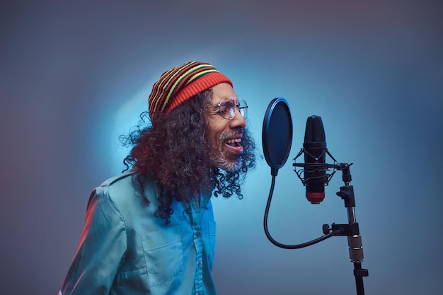 Cantante africano Rastafarian maschio che indossa una camicia blu e un berretto che scrive emotivamente una canzone in studio di registrazione. Isolato su sfondo blu.