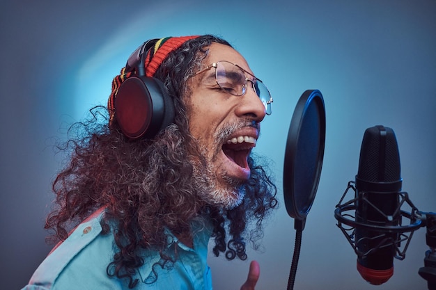 Cantante africano Rastafarian maschio che indossa una camicia blu e un berretto che scrive emotivamente una canzone in studio di registrazione. Isolato su sfondo blu.