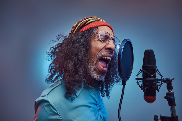 Cantante africano Rastafarian maschio che indossa una camicia blu e un berretto che scrive emotivamente una canzone in studio di registrazione. Isolato su sfondo blu.