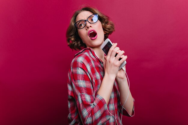 Cantante femminile divertente che scherza sul servizio fotografico al coperto. affascinante signora riccia in bicchieri in posa con smartphone e canto.