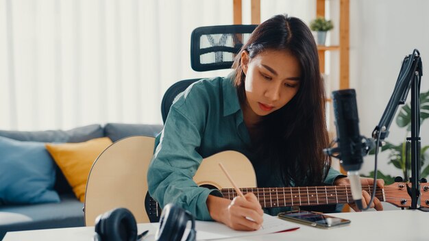 Cantautore donna asiatica felice che suona la chitarra acustica e ascolta la canzone dallo smartphone