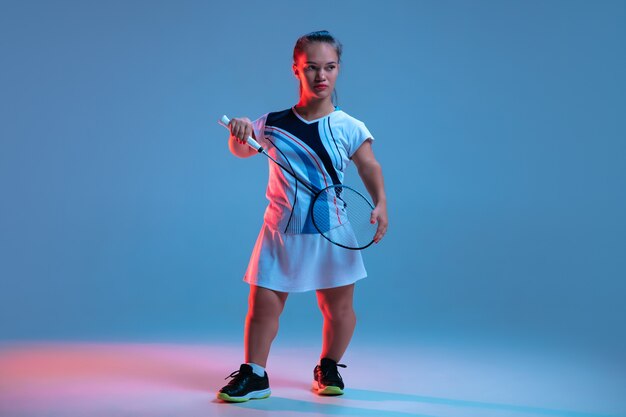 Capo. Bella piccola donna che pratica nel badminton isolata sul blu alla luce al neon. Stile di vita delle persone inclusive, diversità ed uguaglianza