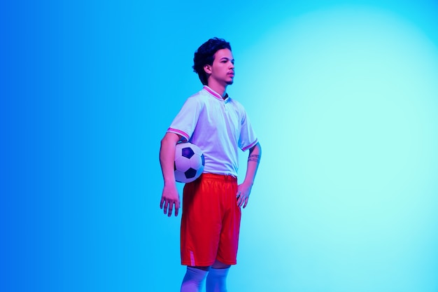 Capo. Giocatore di calcio o di calcio sulla parete dello studio blu sfumato alla luce al neon - posa sicura con la palla. Copyspace.