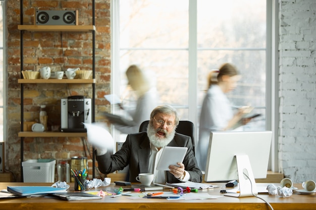 Capo nervoso e stanco al suo posto di lavoro occupato mentre le persone si muovono vicino sfocate. Impiegato, manager che lavora, ha problemi e scadenze, i suoi colleghi distraggono. Affari, lavoro, concetto di carico di lavoro.