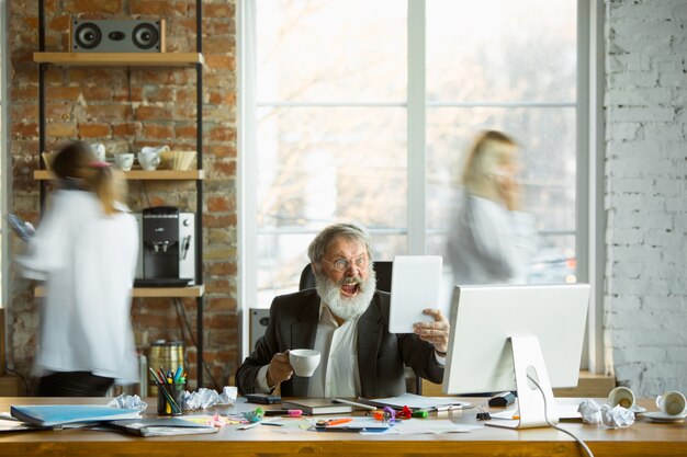 Capo nervoso e stanco al suo posto di lavoro occupato mentre le persone si muovono vicino sfocate. Impiegato, manager che lavora, ha problemi e scadenze, i suoi colleghi distraggono. Affari, lavoro, concetto di carico di lavoro.