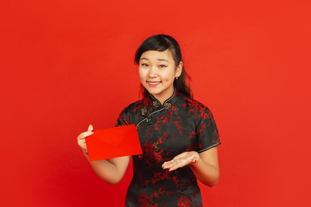 Capodanno cinese. Ritratto di ragazza asiatica isolato su sfondo rosso. Modello femminile in abiti tradizionali sembra felice, sorridente e mostrando la busta rossa. Celebrazione, vacanza, emozioni.
