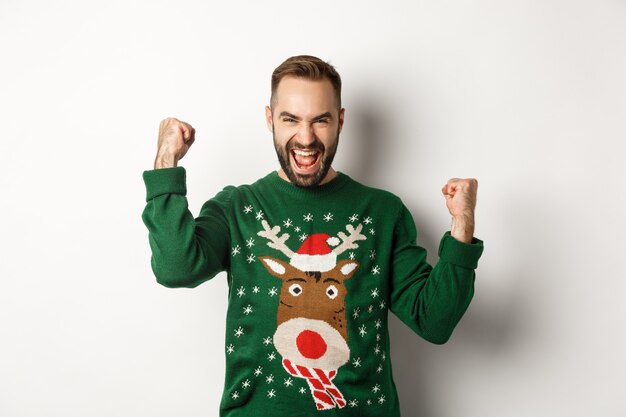 Capodanno, vacanze e celebrazione. Eccitato uomo barbuto con un maglione natalizio, che fa pompe a pugno e grida di gioia, rallegrandosi e trionfando, sfondo bianco.