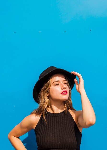 Cappello d&#39;uso della giovane donna alla moda che sta davanti a fondo blu