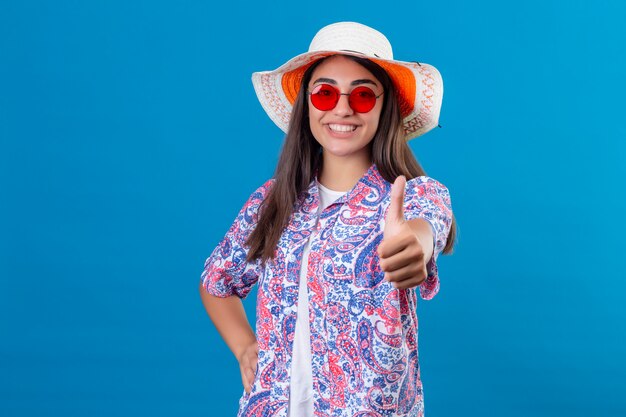 Cappello d'uso di estate del giovane bello turista della donna e occhiali da sole rossi che sorridono con il fronte felice che mostra i pollici su sopra la parete blu isolata