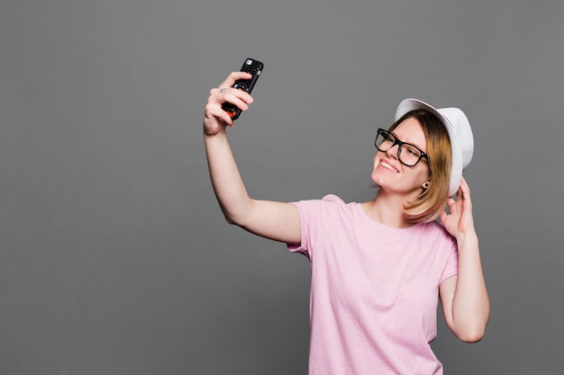 Cappello d&#39;uso sorridente della giovane donna che prende selfie sul telefono cellulare contro il contesto grigio