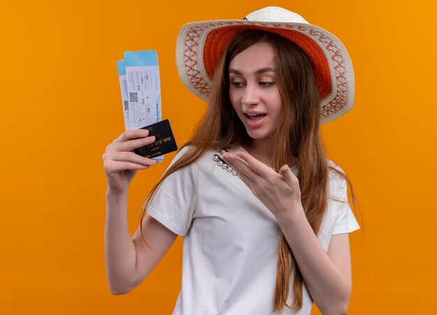 Cappello da portare impressionato della ragazza del giovane viaggiatore che tiene i biglietti aerei, carta di credito che indica con la mano su uno spazio arancione isolato
