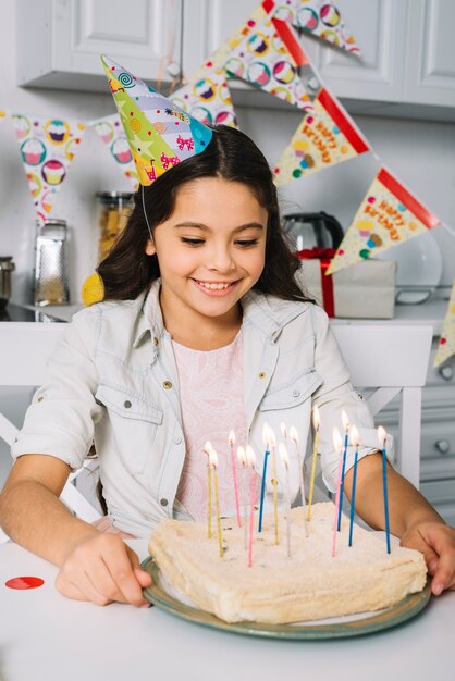 Cappello di partito sorridente della ragazza di compleanno che porta sulla testa che esamina dolce decorato con le candele variopinte