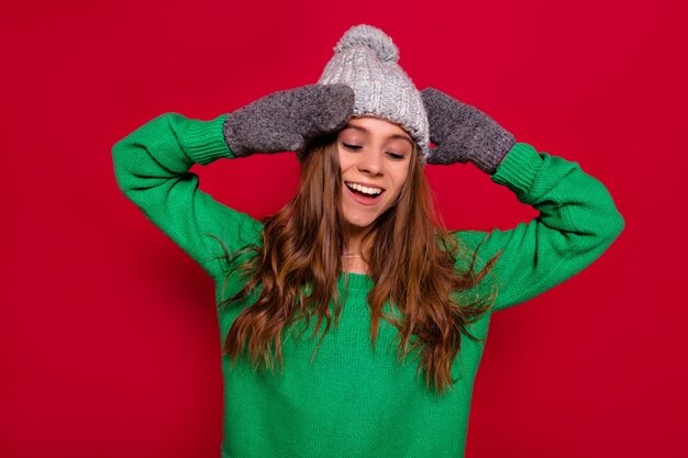 Carina bella donna felice con capelli lunghi vestita berretto invernale e maglione verde in piedi su sfondo rosso con gli occhi chiusi, sorriso felice ed emozioni calme