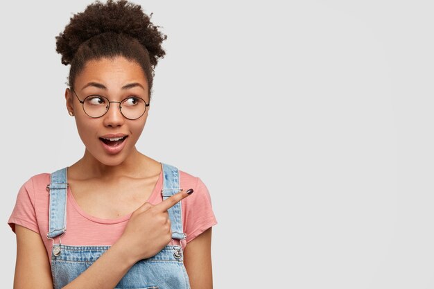 Carina bella giovane femmina dalla pelle scura con capelli ricci, ha una grande idea, indica con il dito anteriore a parte, vestita casualmente, isolata sopra il muro bianco Persone, sorpresa e concetto di shock