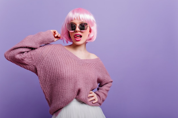 Carina donna europea in periwig colorato in posa con la lingua fuori. fiduciosa ragazza beata con i capelli rosa con gli occhiali e maglione viola.