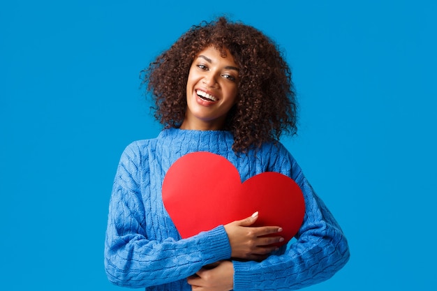 Carina e tenera, divertente, sorridente femmina afro-americana con taglio di capelli afro, premere il grande segno di cuore rosso sul petto e abbracciarlo con un sorriso affascinante e felice, mostrando amore e affetto, muro blu.