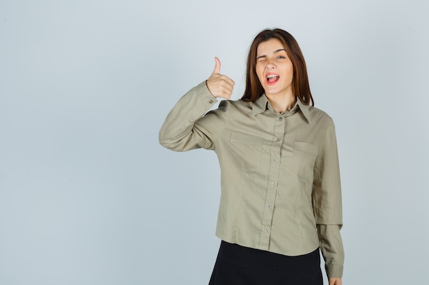 Carina giovane donna che mostra pollice in su mentre sbatte le palpebre in camicia, gonna e sembra orgogliosa, vista frontale.