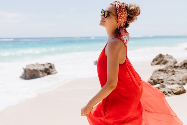 Carina giovane donna con la pelle leggermente abbronzata in posa con il sorriso in riva al mare. Affascinante modello femminile agghiacciante sulla natura in una giornata di sole.