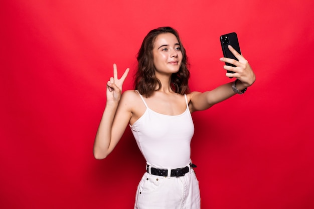 Carina giovane donna divertendosi e prendendo selfie con gesti segno di pace isolato su rosso