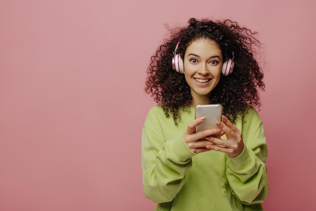 Carina giovane ragazza bruna caucasica in cuffia guarda la fotocamera su sfondo rosa Bellezza riccia in giacca verde tiene il telefono nelle sue mani Concetto di tecnologia e persone