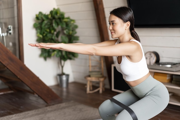 Carina ragazza asiatica di fitness a casa che fa squat di allenamento con una corda elastica allungata sulle gambe eserci...