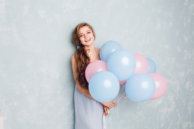 Carina ragazza bionda in piedi in uno studio, sorridente e con palloncini blu e rosa.