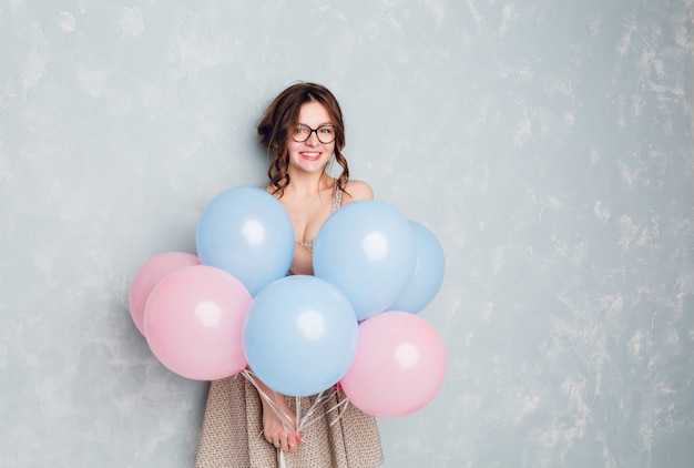 Carina ragazza bruna in piedi in uno studio, sorridente ampiamente e con palloncini blu e rosa.